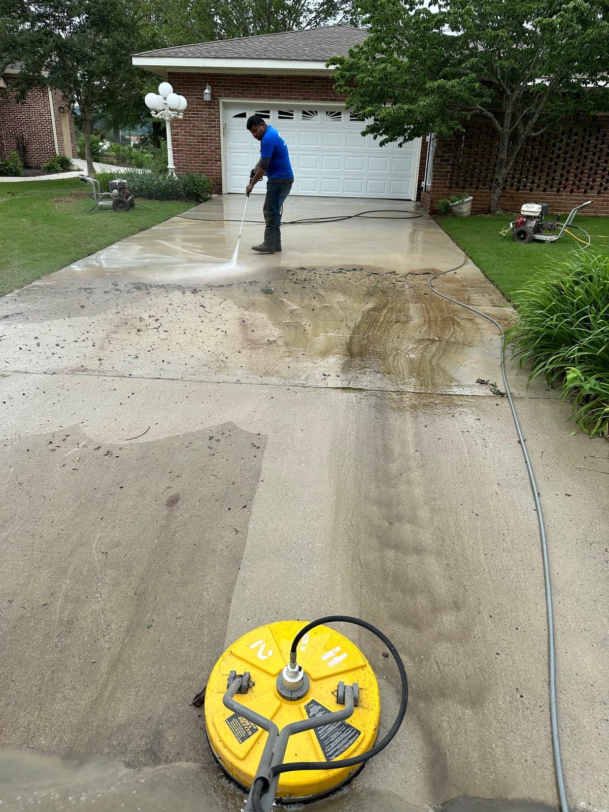 A crew member washing a concrete driveway with a pressure wand, the cleaned strip showing bright against the dirty concrete.